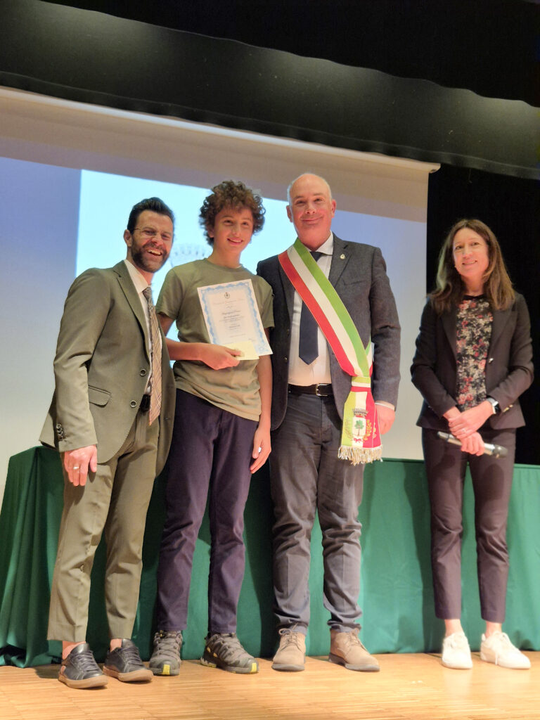 Un giovane studente sul palco sorride mentre mostra un attestato di merito consegnato dal sindaco con la fascia tricolore e da altri rappresentanti istituzionali.