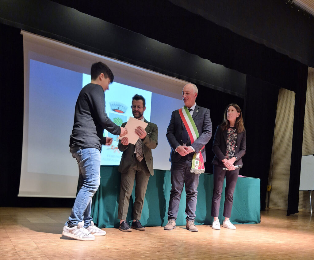 Cerimonia di premiazione su un palco comunale. Un giovane studente in jeans riceve un certificato da un assessore, affiancato dal sindaco con la fascia tricolore italiana. Una presentatrice con microfono è a lato. Sullo sfondo, un logo comunale proiettato.